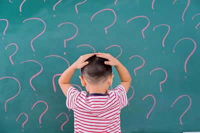 A boy holds his hands on his head while looking at a blackboard filled with question marks.